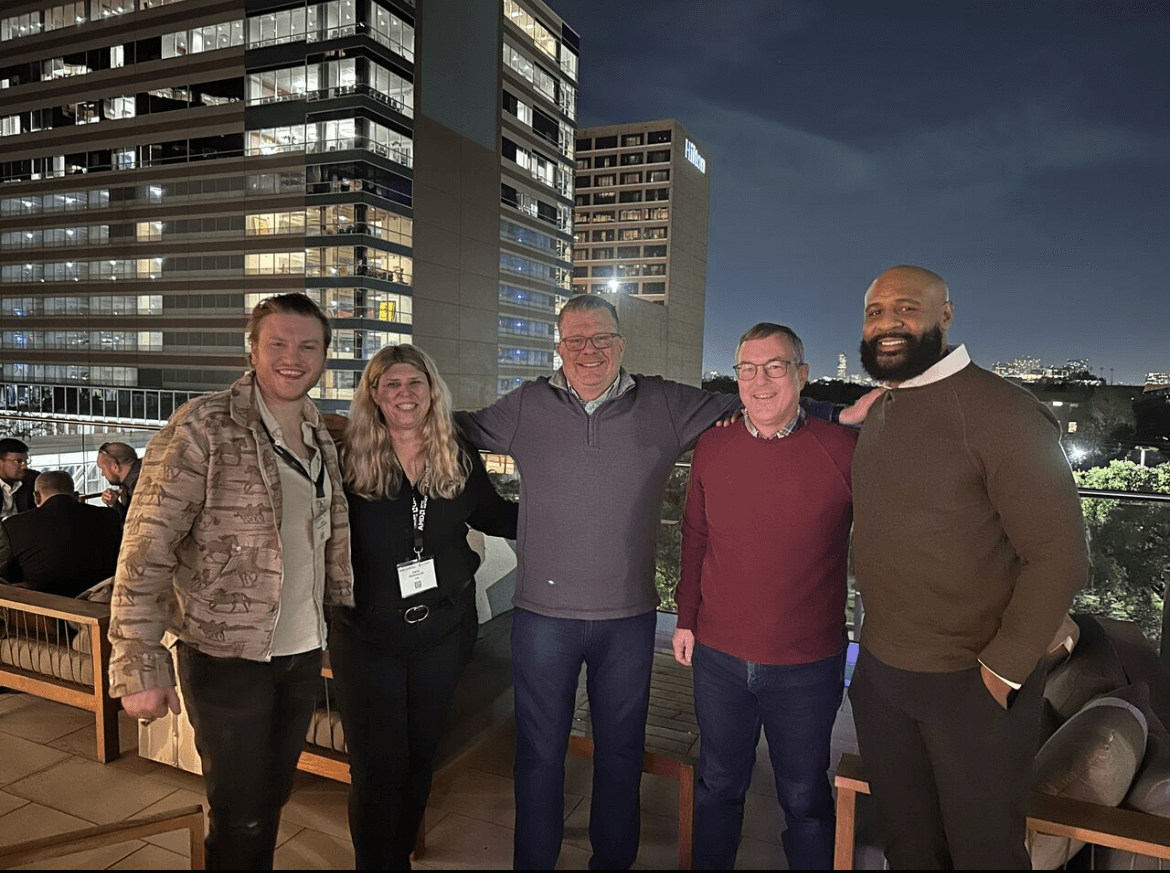 Five Vultr employees standing together on a rooftop terrace at night with illuminated office buildings in the background. They're smiling with arms around each other's shoulders.